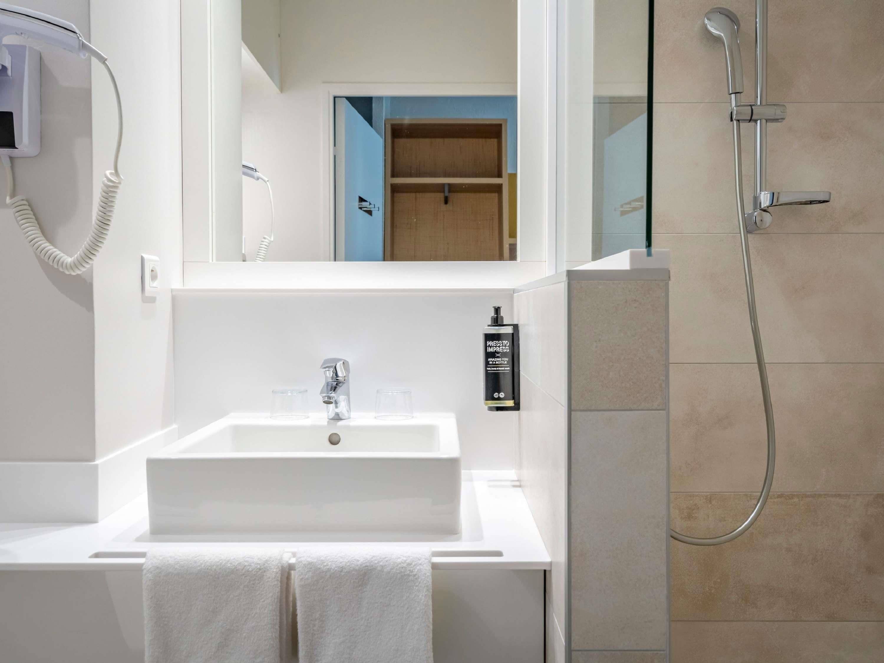 A modern bathroom with a white sink, a mirror, a handheld showerhead, and a bottle of press to impress.