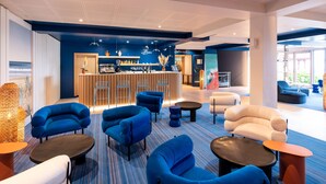 A modern lounge area with blue and white seating, a bar with a wooden counter, and a large window offering a view of the outdoors. - ibis Styles Biarritz Plage (Ouverture mai 2024) (Biarritz)