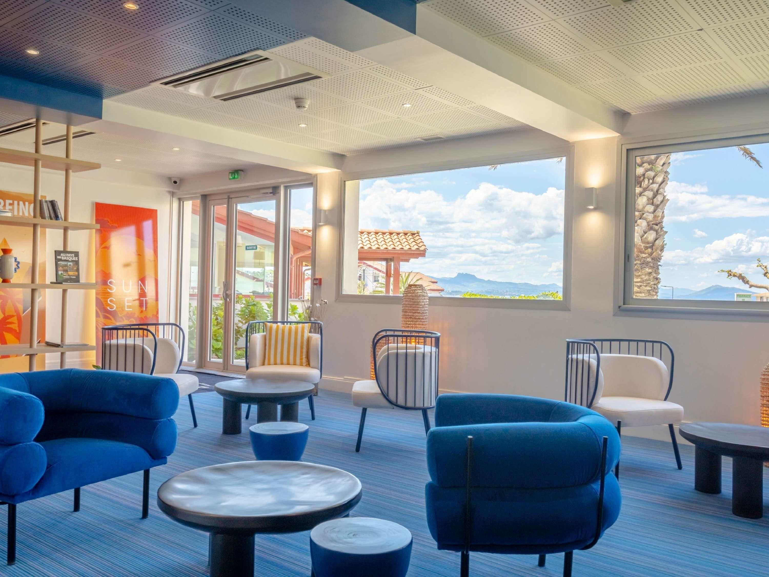 Un espace de détente moderne avec des fauteuils bleus, des tables rondes et une vue sur l’extérieur.