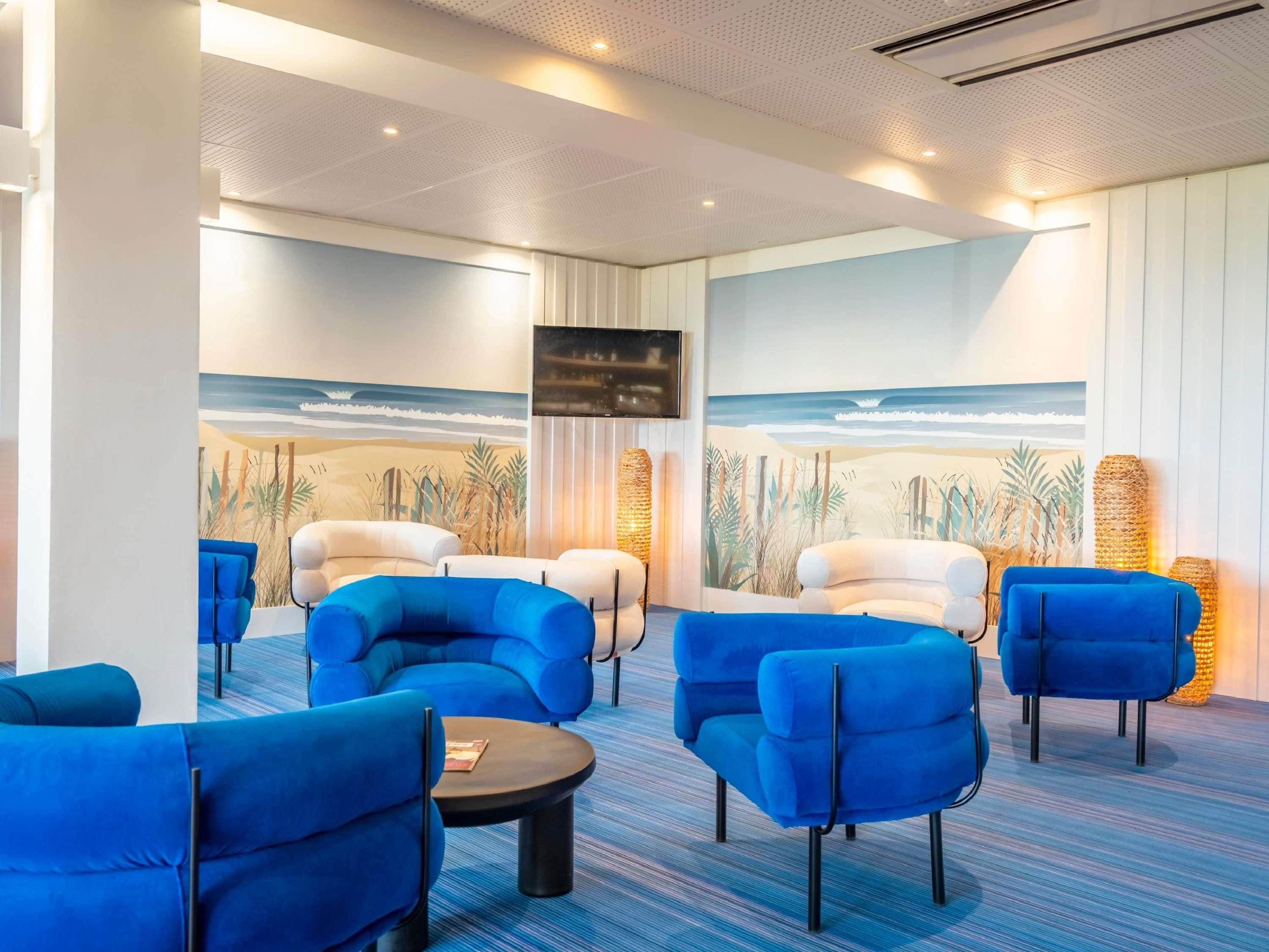 A modern lounge area with blue upholstered chairs, a round table, and a beach mural on the wall.