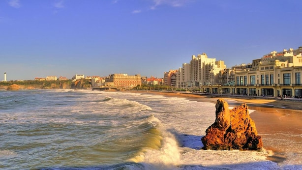 A coastal cityscape with a prominent lighthouse, beachfront buildings, and a rocky outcrop in the foreground. - Le Petit Hôtel (Biarritz)