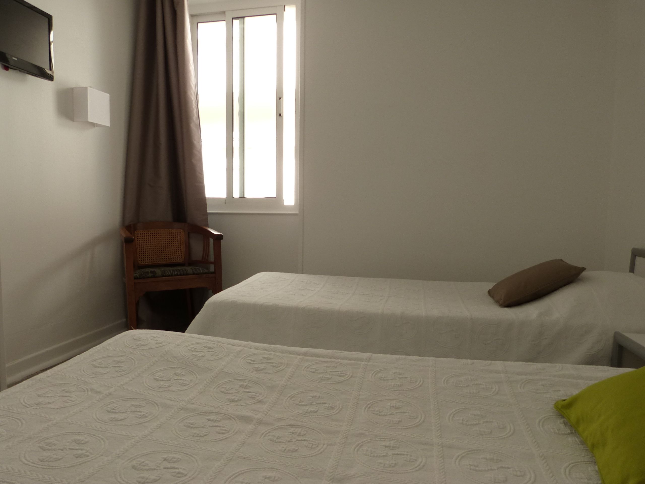 A room with two beds, a chair, a window with curtains, and a flat-screen TV mounted on the wall.