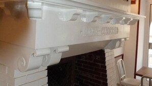 A fireplace with a white mantel and decorative moldings, flanked by framed wall art and a brick wall below. - Le Petit Hôtel (Biarritz)
