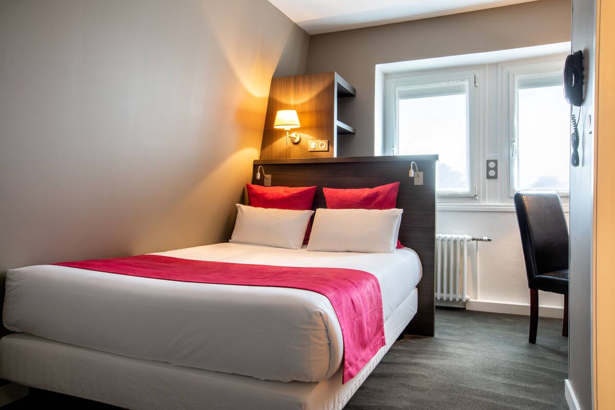 Ein Hotelzimmer mit Bett, Nachttisch, Lampe, Stuhl und Fenster.