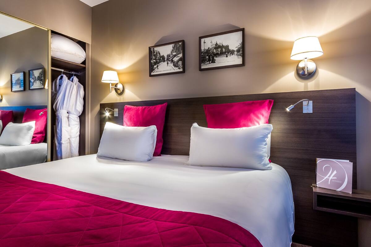 Ein Hotelzimmer mit einem großen Bett, rot-weißer Bettwäsche, einem Holz-Kopfteil, zwei Wandleuchten, einem Spiegel und zwei gerahmten Bildern.