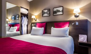 Une chambre d’hôtel avec un grand lit, une literie rouge et blanche, une tête de lit en bois, deux lampes fixées au mur, un miroir et deux tableaux encadrés.