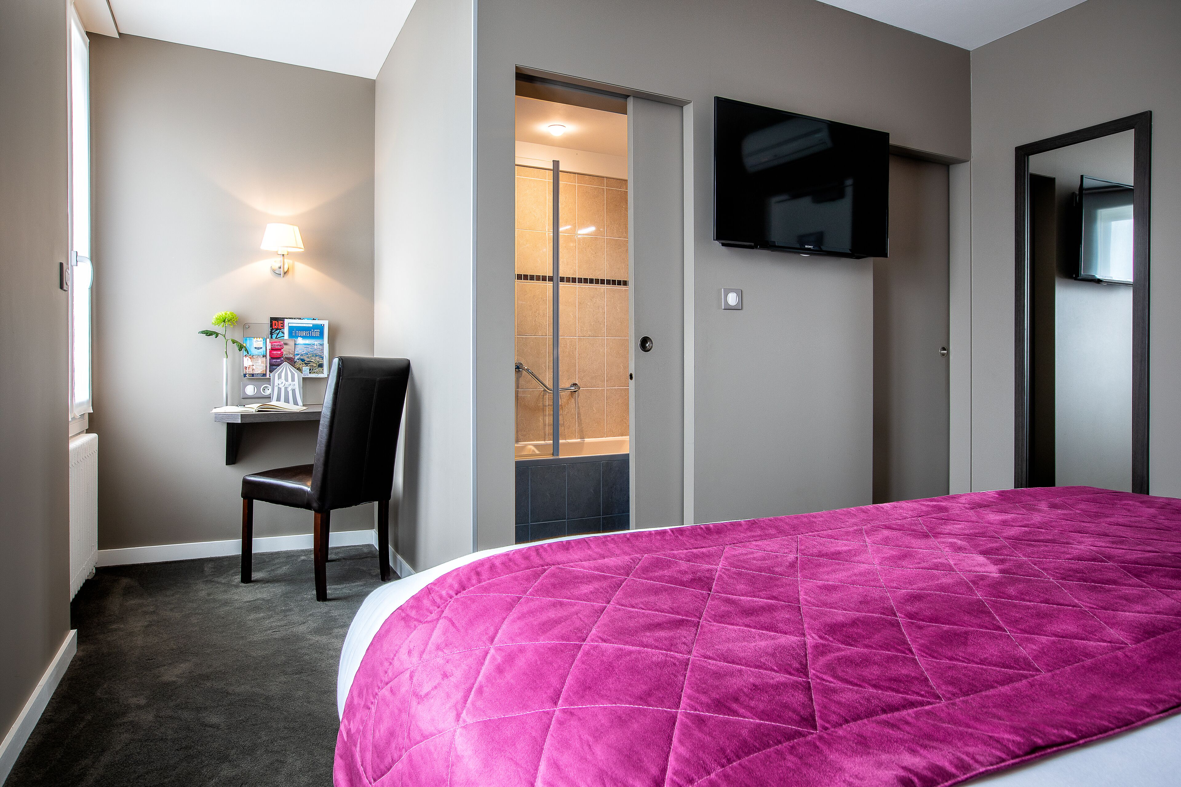A hotel room with a bed, a desk with a chair, a TV, and a shower area.