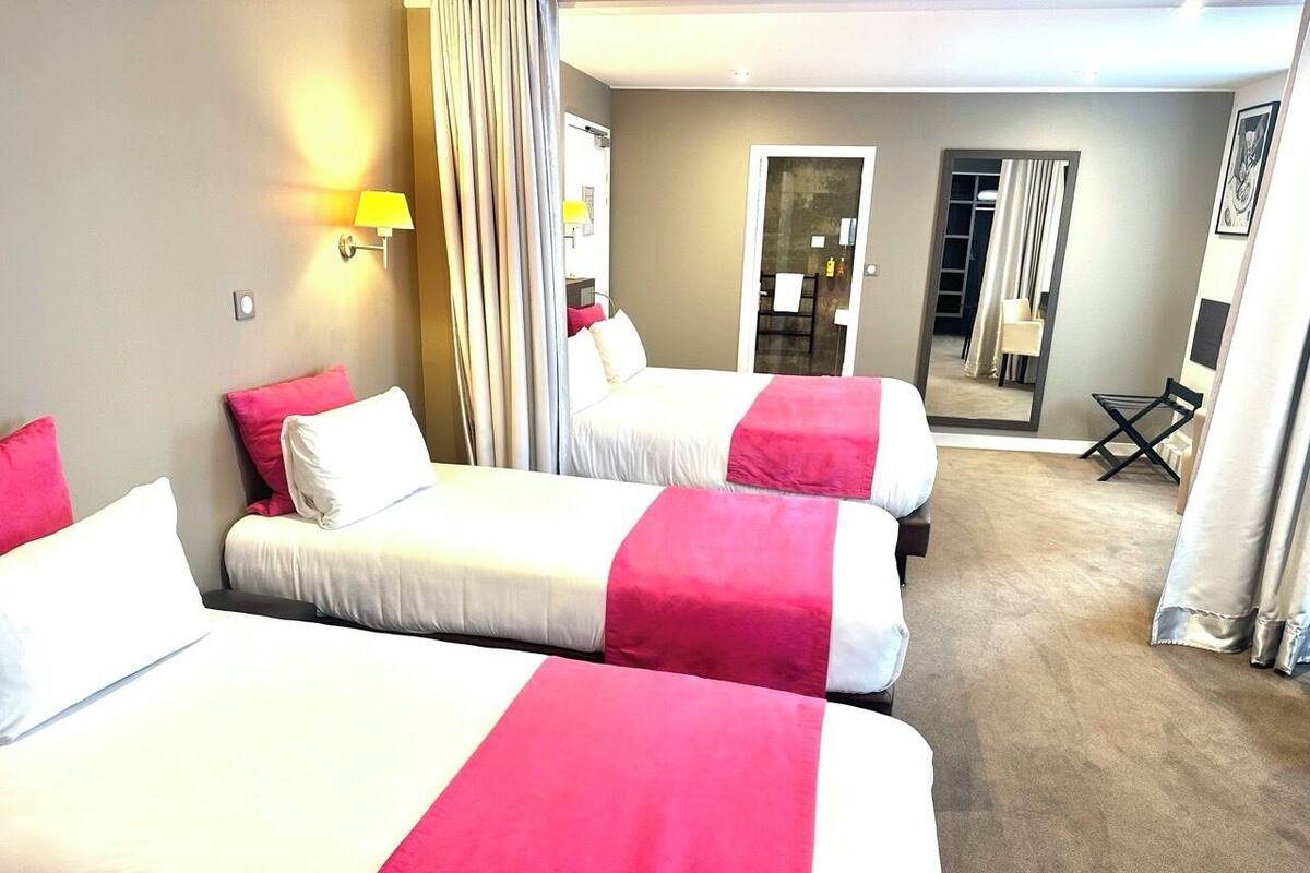 Ein Hotelzimmer mit drei Betten, jeweils mit einer rosa und weißen Tagesdecke, einem kleinen Tisch und einem Stuhl. An der Wand befindet sich ein Spiegel und eine Tür zu einem Badezimmer.
