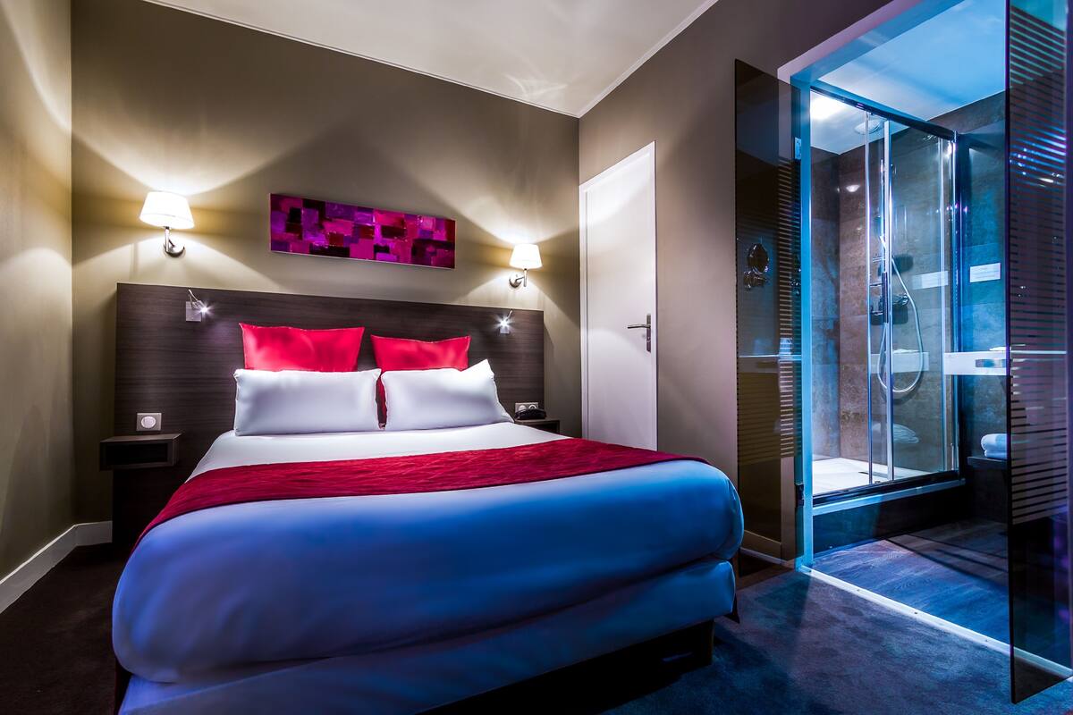 Ein modernes Hotelzimmer mit einem großen Bett, zwei Nachttischen, einer gläsernen Dusche und einem an der Wand befestigten Kunstwerk.