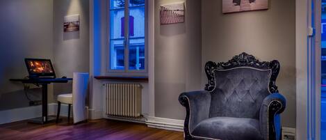 A room with a large window, a dark upholstered armchair, a small table with a chair, and artwork on the walls.