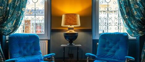A room with two blue armchairs, a glass table, a lamp, and a view through a window with curtains.