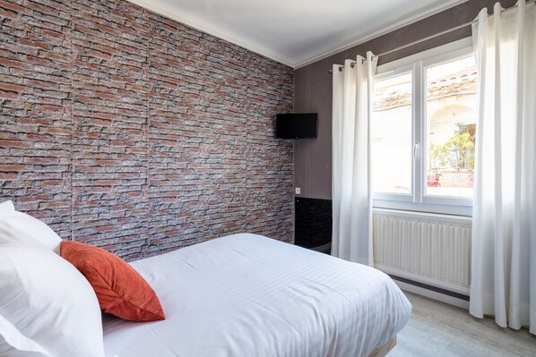 A bedroom with a brick wall, a bed with white and orange pillows, a mounted TV, and a window with curtains. - La Maison d'été (Salon-de-Provence)