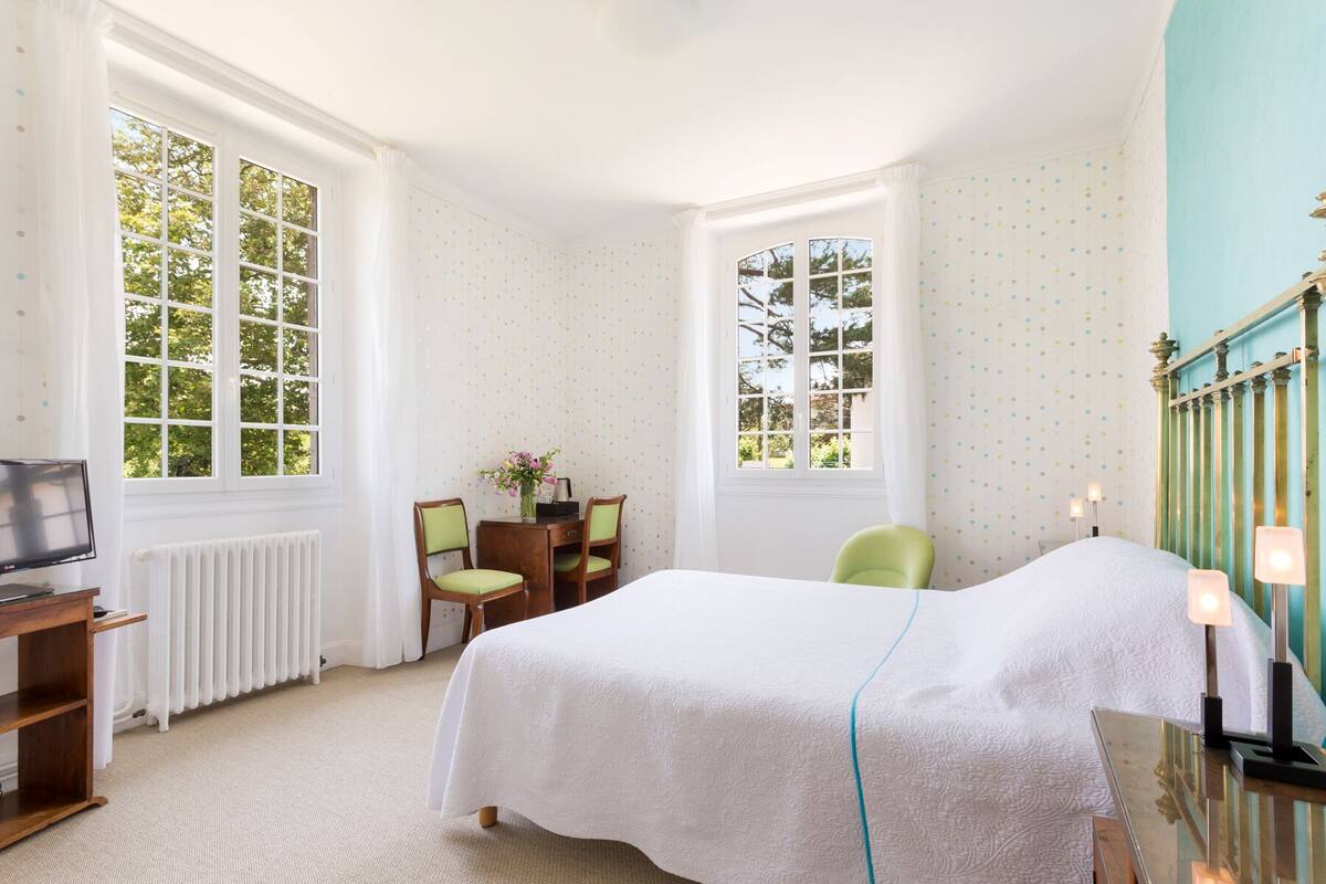 Un dormitorio con cama, escritorio, dos sillas, radiador y ventanas amplias.