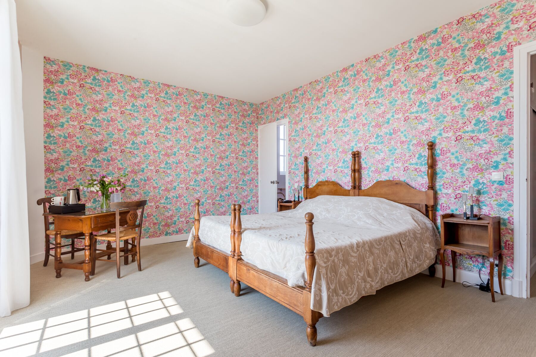 Un dormitorio con papel tapiz floral, una cama de madera, un escritorio con silla y una mesita de noche.