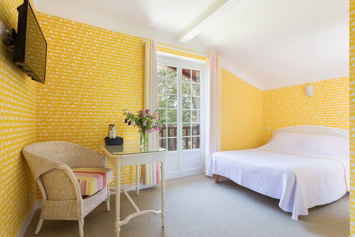 Una habitación con papel tapiz amarillo estampado, una cama, un sillón de mimbre, una mesita con un jarrón de flores y una ventana con vistas a árboles.