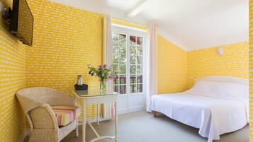 A room with yellow patterned wallpaper, a bed, a wicker chair, a small table with a vase of flowers, and a window with a view of trees.