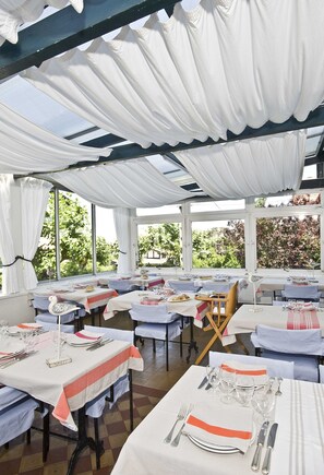 A dining area with tables set for multiple guests, large windows, and a view of greenery outside. - Hotel Les Goelands (Saint-Jean-de-Luz)