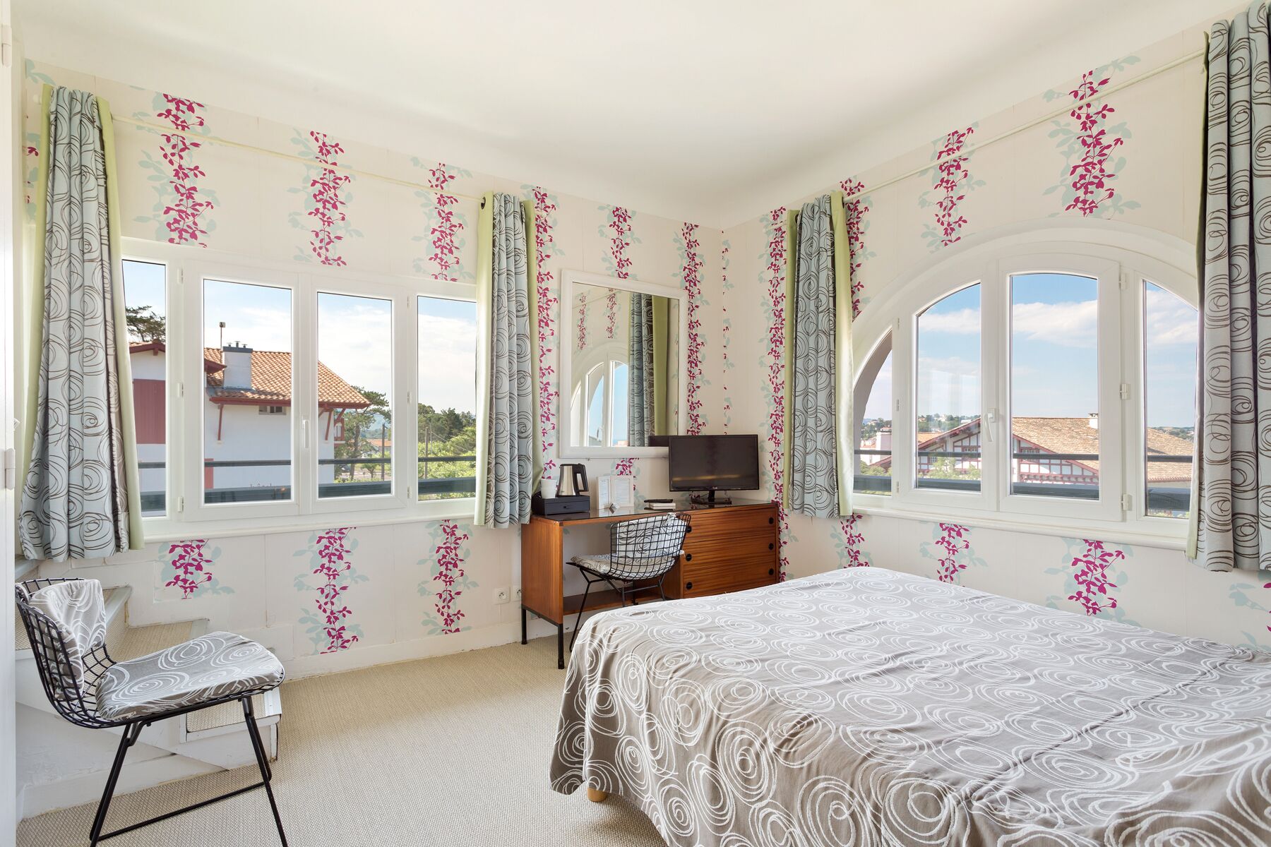 A bedroom with a bed, a desk with a chair, a TV, and large windows with curtains.