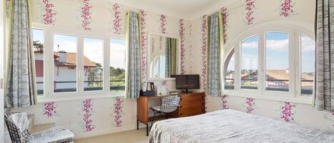 A bedroom with a bed, a desk with a chair, a TV, and large windows with curtains.