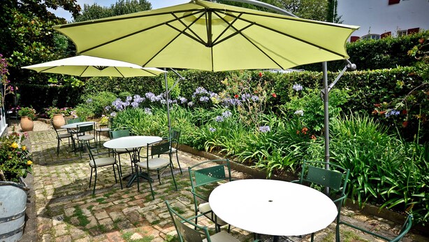 Une terrasse extérieure avec des chaises et des tables en métal vert, entourée de parterres de fleurs luxuriantes et d’une haie.