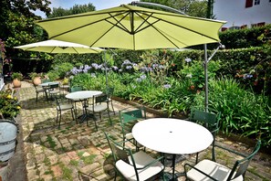 Een terras buiten met groene metalen stoelen en tafels, omringd door weelderige bloembedden en een haag.