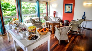 Un buffet de petit-déjeuner proposant une variété d’aliments et de boissons, des meubles en osier et de grandes fenêtres avec des rideaux.