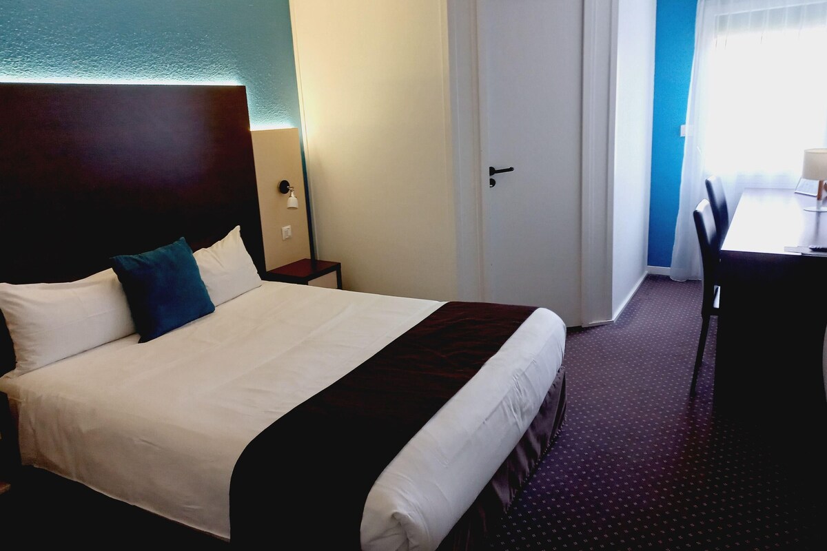 A hotel room with a large bed, a bedside table, a desk with a chair, and a door.