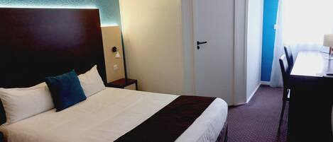 A hotel room with a large bed, a bedside table, a desk with a chair, and a door.