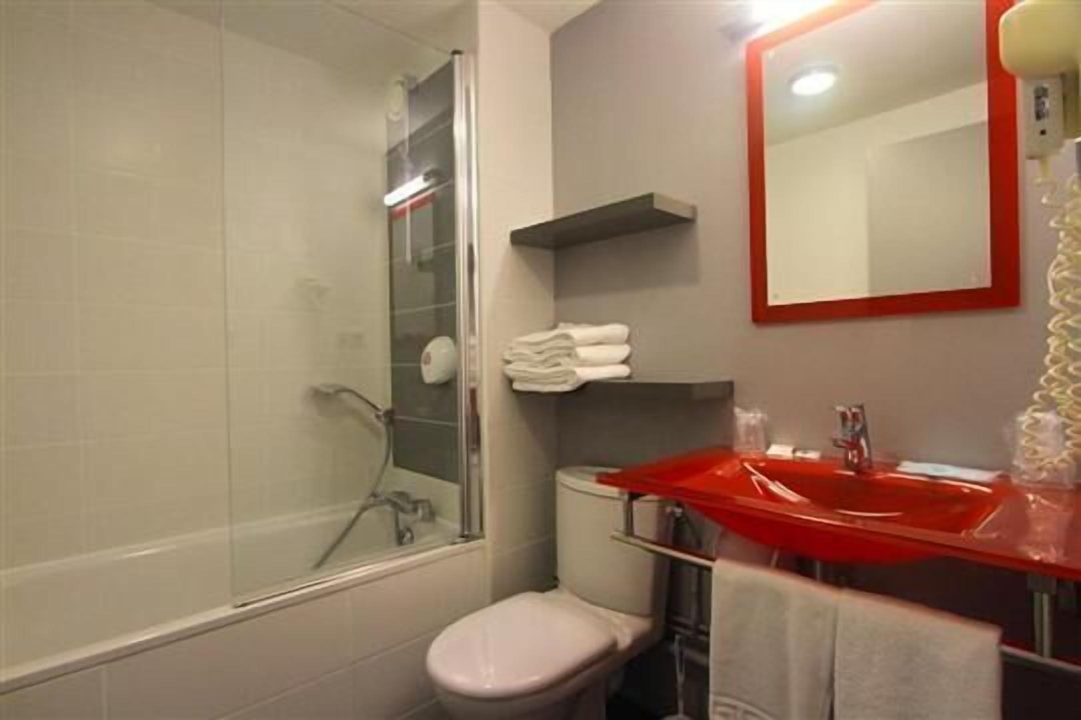 A bathroom with a glass shower enclosure, a white toilet, a red sink, and a mirror with a red frame.
