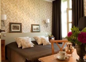 Habitación de hotel con cama, dos cuadros en la pared, una mesa con taza y plato, y un florero con flores.