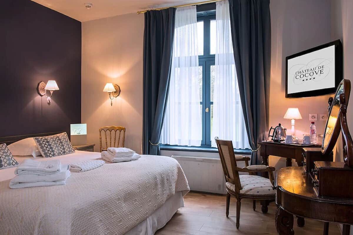 A hotel room with a bed, bedside lamps, a piano, a desk with a lamp, and a window with curtains.