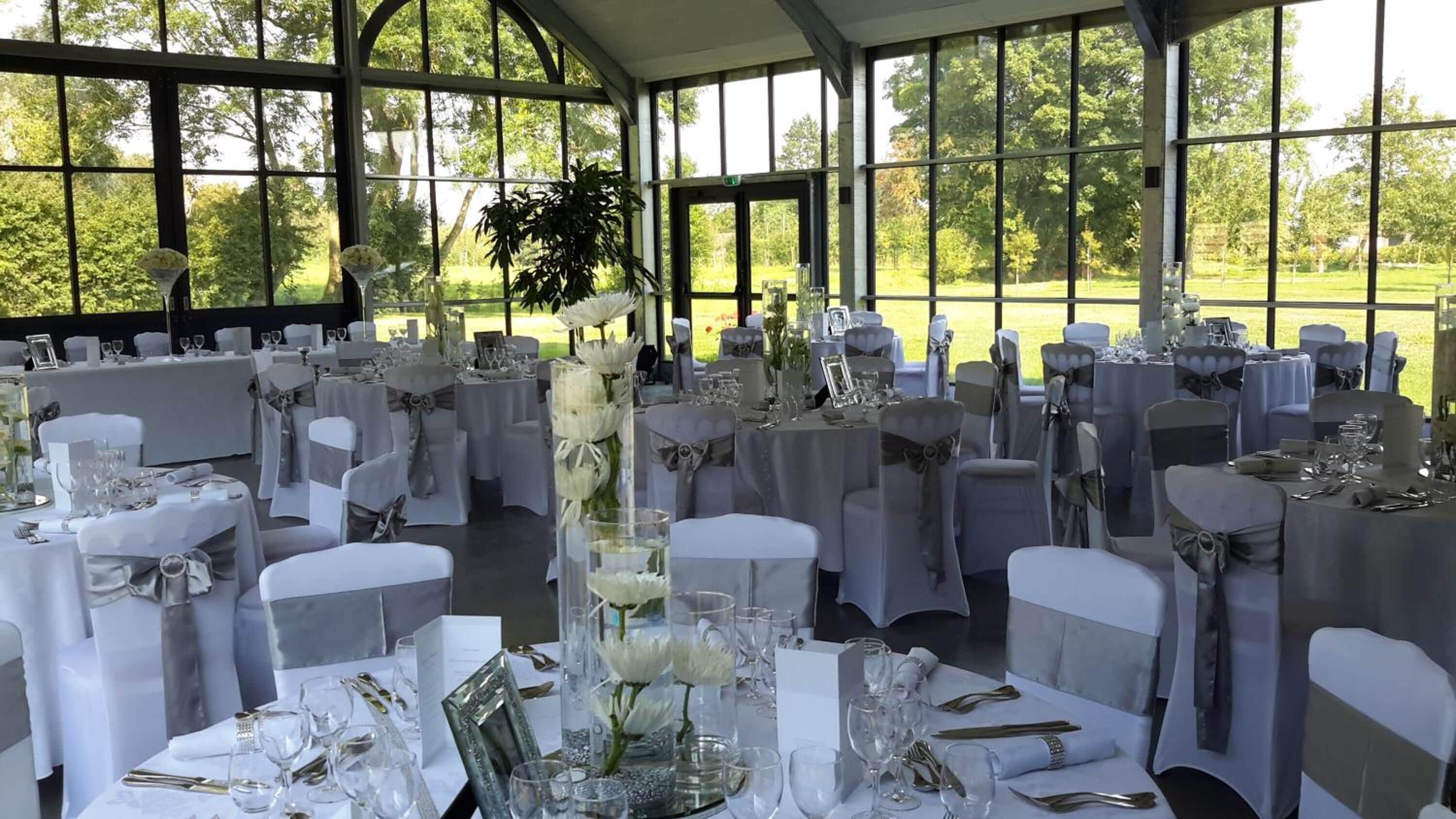 Une salle de banquet avec des tables rondes dressées pour un événement formel, agrémentée de nappes blanches, de couverts en argent et de verres.
