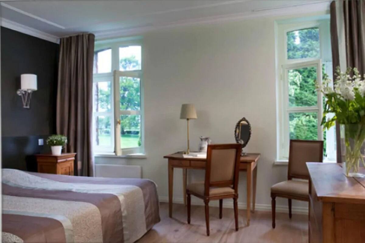 A hotel room with a bed, a desk with a chair, a lamp, and a window with curtains.