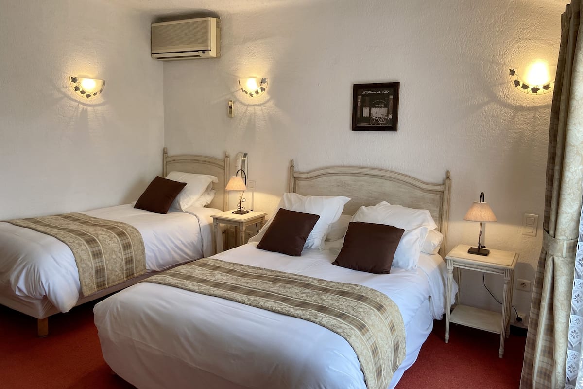Une chambre d’hôtel avec deux lits simples, chacun doté d’une table de chevet et d’une lampe, un grand lit recouvert d’une couverture à motifs, un climatiseur fixé au mur et un tableau encadré accroché au mur.