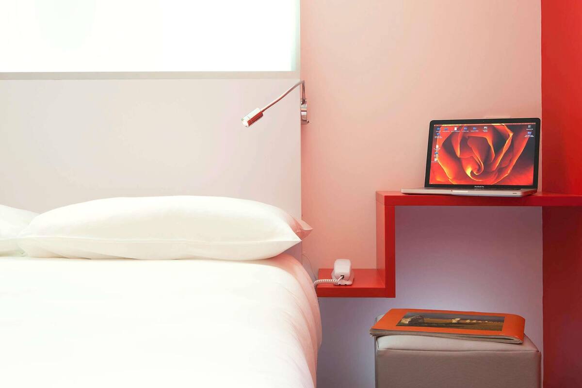 Un lit bien fait, avec des oreillers blancs et une table de chevet rouge sur laquelle se trouvent un ordinateur portable et un livre.