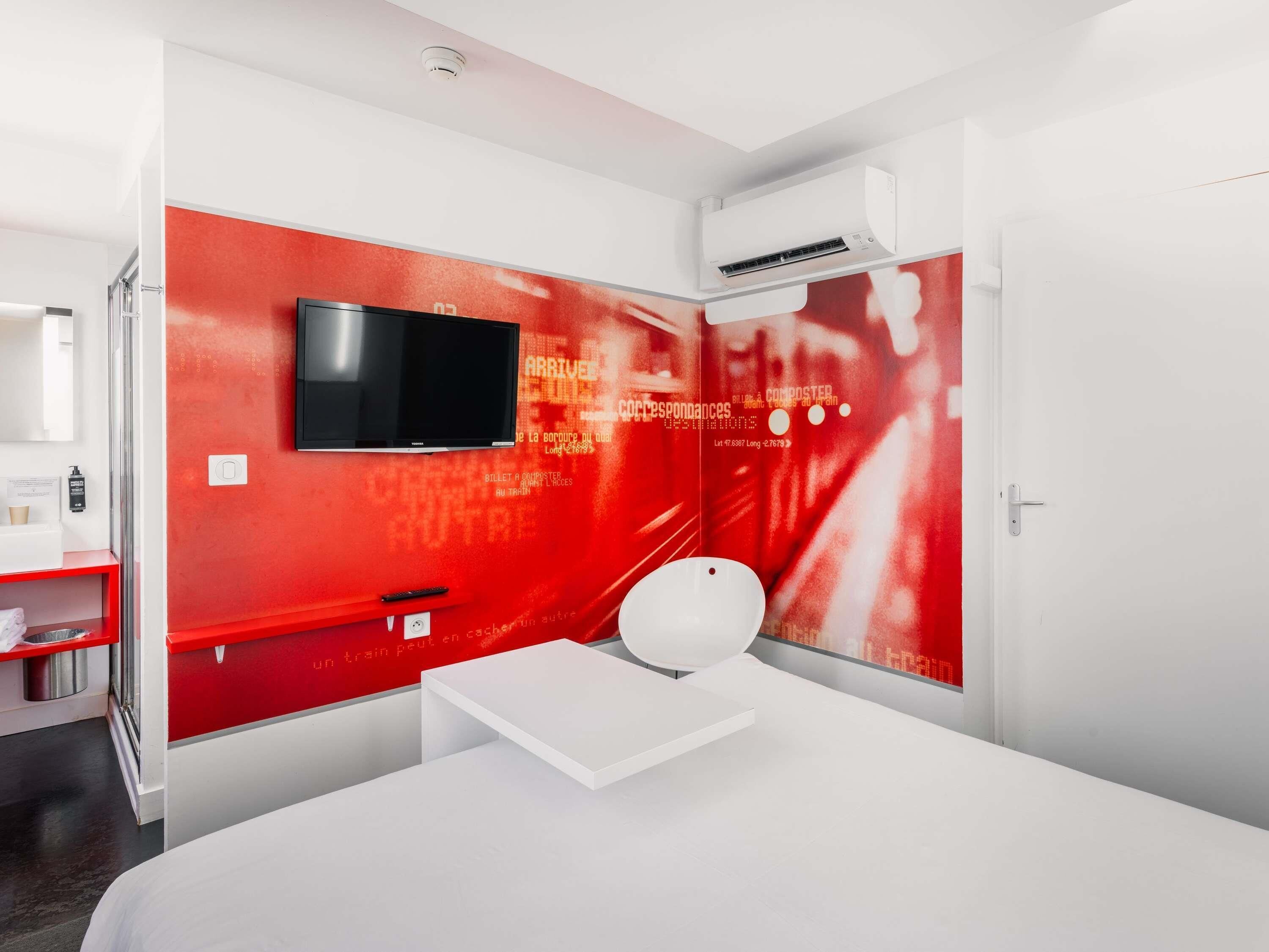 A modern hotel room with a red accent wall, a mounted television, a white bedside table, and a white toilet.