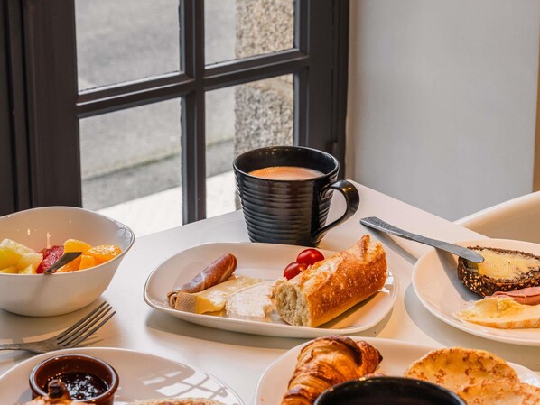 Una mesa de desayuno con un tazón de fruta, croissants, medio croissant, una rebanada de pan con queso, y una taza de café.