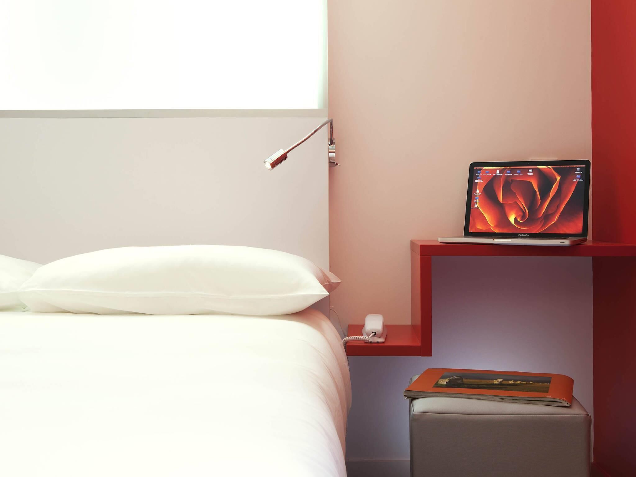 Une chambre d’hôtel avec un lit, une table de chevet avec une lampe, un ordinateur portable et une petite étagère avec un livre.