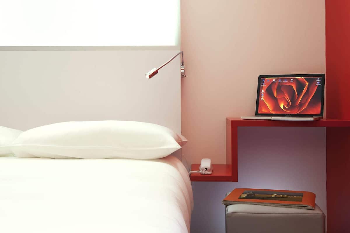 A hotel room with a bed, a bedside table with a lamp, a laptop, and a small shelf with a book.