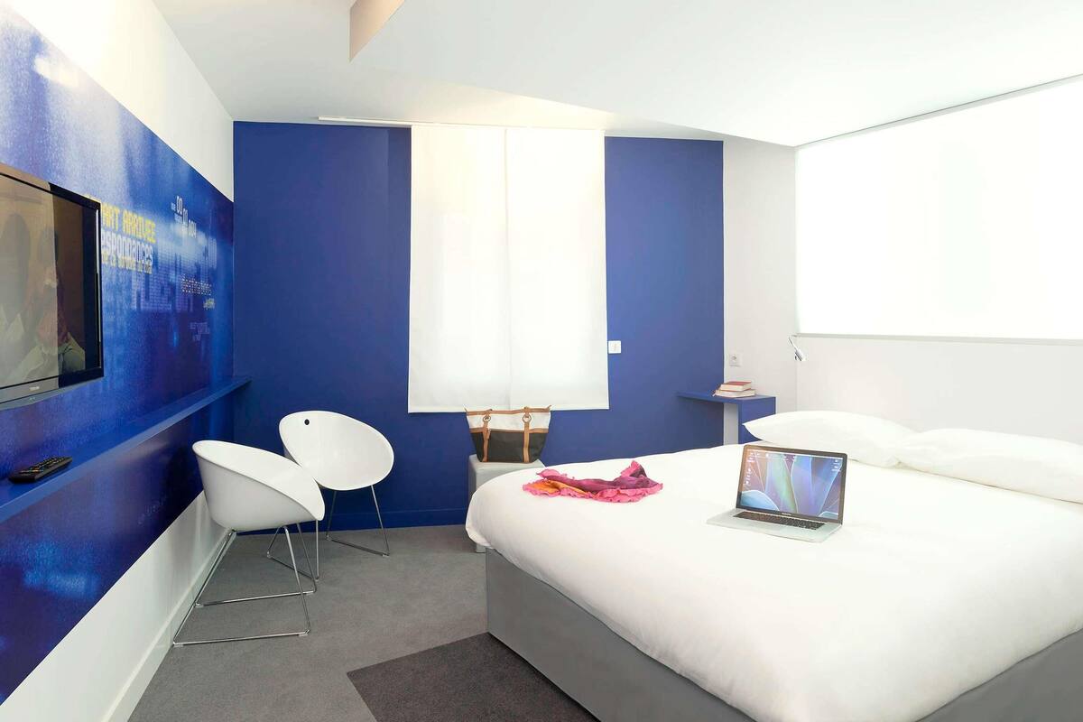 Een moderne hotelkamer met een groot bed, een bureau, twee witte stoelen, een televisie aan de muur en een laptop op het bed.