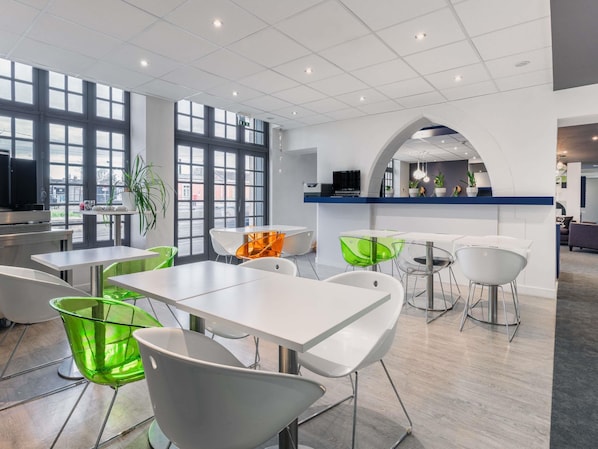 A modern dining area with white tables and chairs, large windows, and a kitchen area in the background. - ibis Styles Vannes Gare Centre (Vannes)