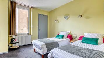 A hotel room with two beds, a small table, a radiator, a window with curtains, and a door.
