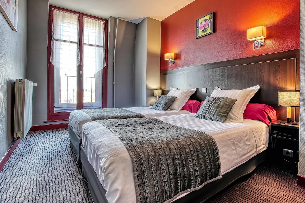 Ein Hotelzimmer mit zwei Betten, einer roten Akzentwand, einem großen Fenster mit Vorhängen und einem Nachttisch mit Lampe.