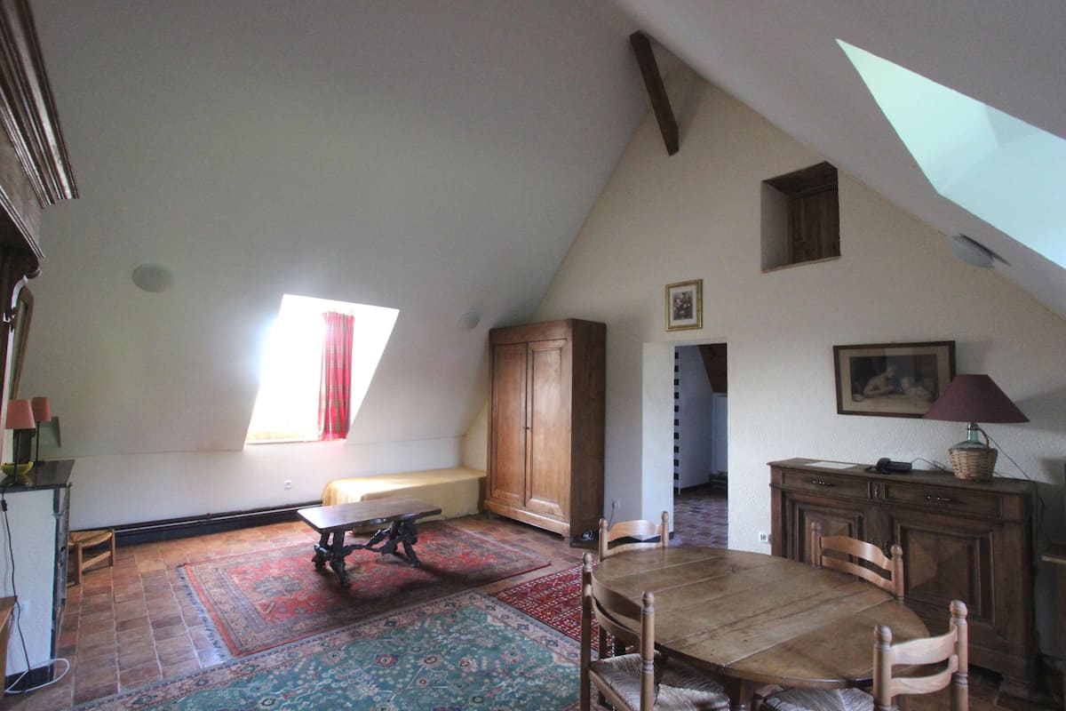 Une vaste pièce mansardée comprenant une grande table à manger en bois, un banc et une vue sur le ciel grâce à une lucarne.