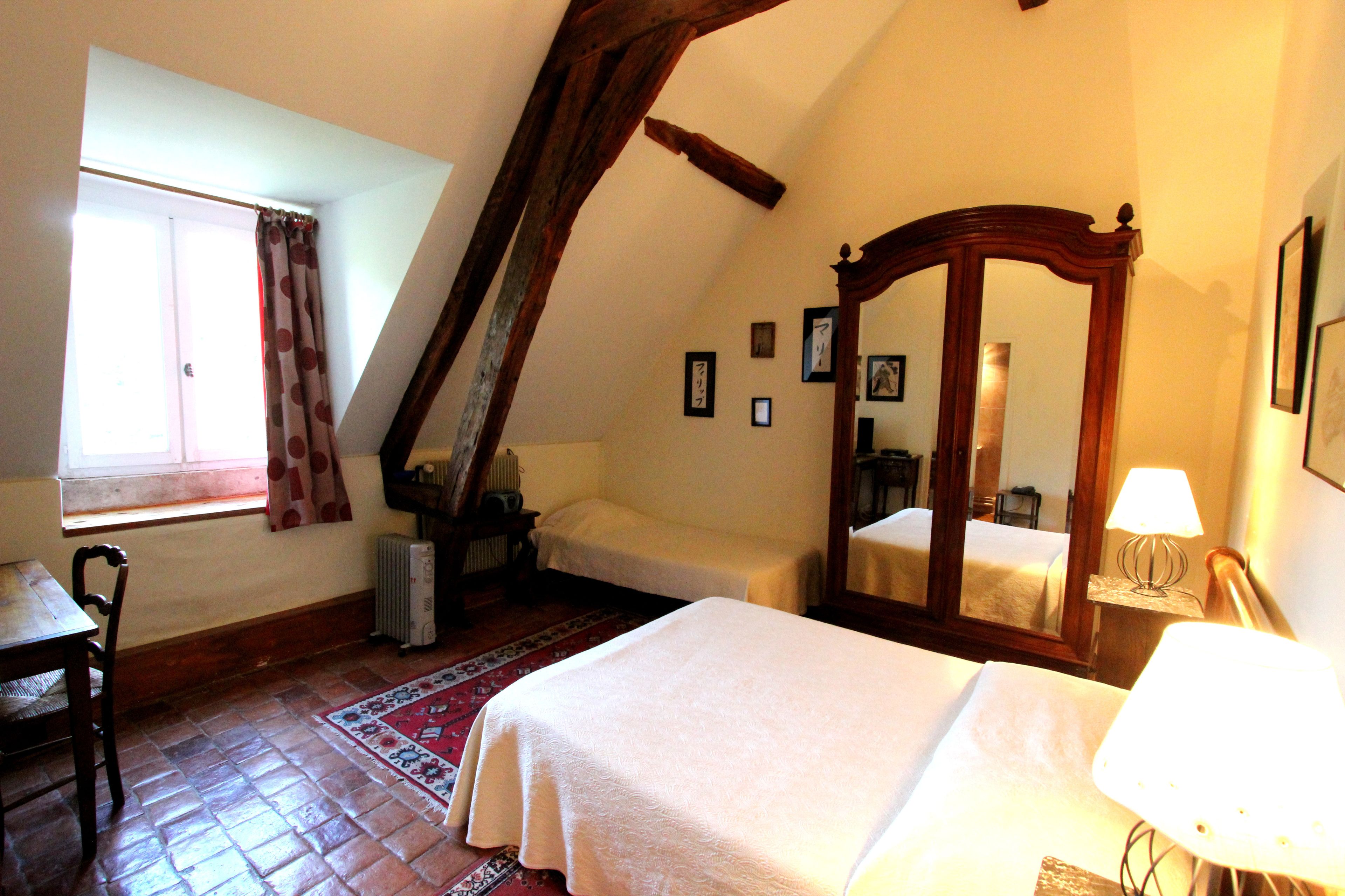 A bedroom with a large bed, a chair, a desk, a window with curtains, a radiator, a mirror, and framed pictures on the wall.