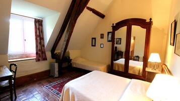 A bedroom with a large bed, a chair, a desk, a window with curtains, a radiator, a mirror, and framed pictures on the wall.