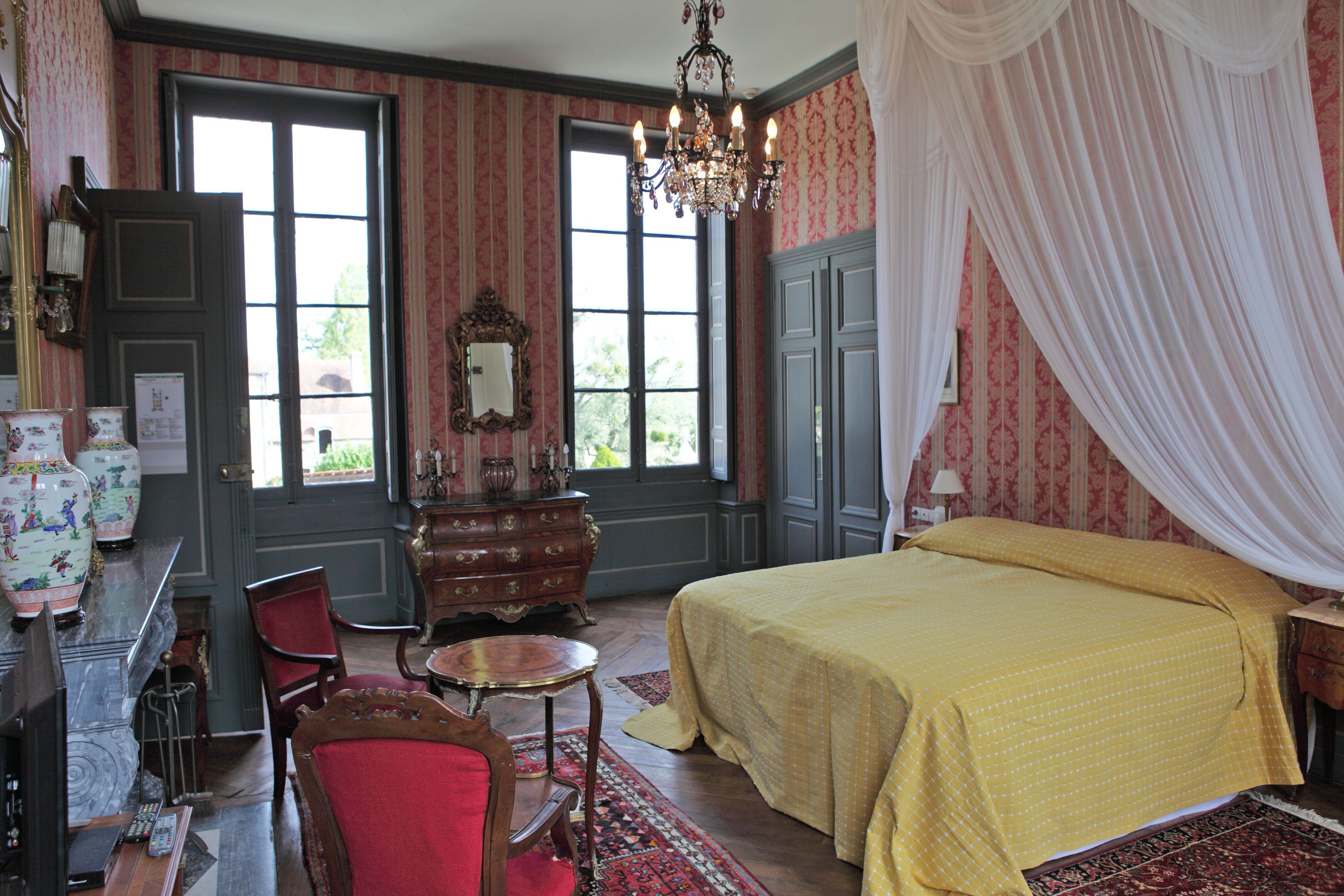 Una habitación con una cama grande, una araña, una silla roja, un tocador de madera y una ventana con cortinas.
