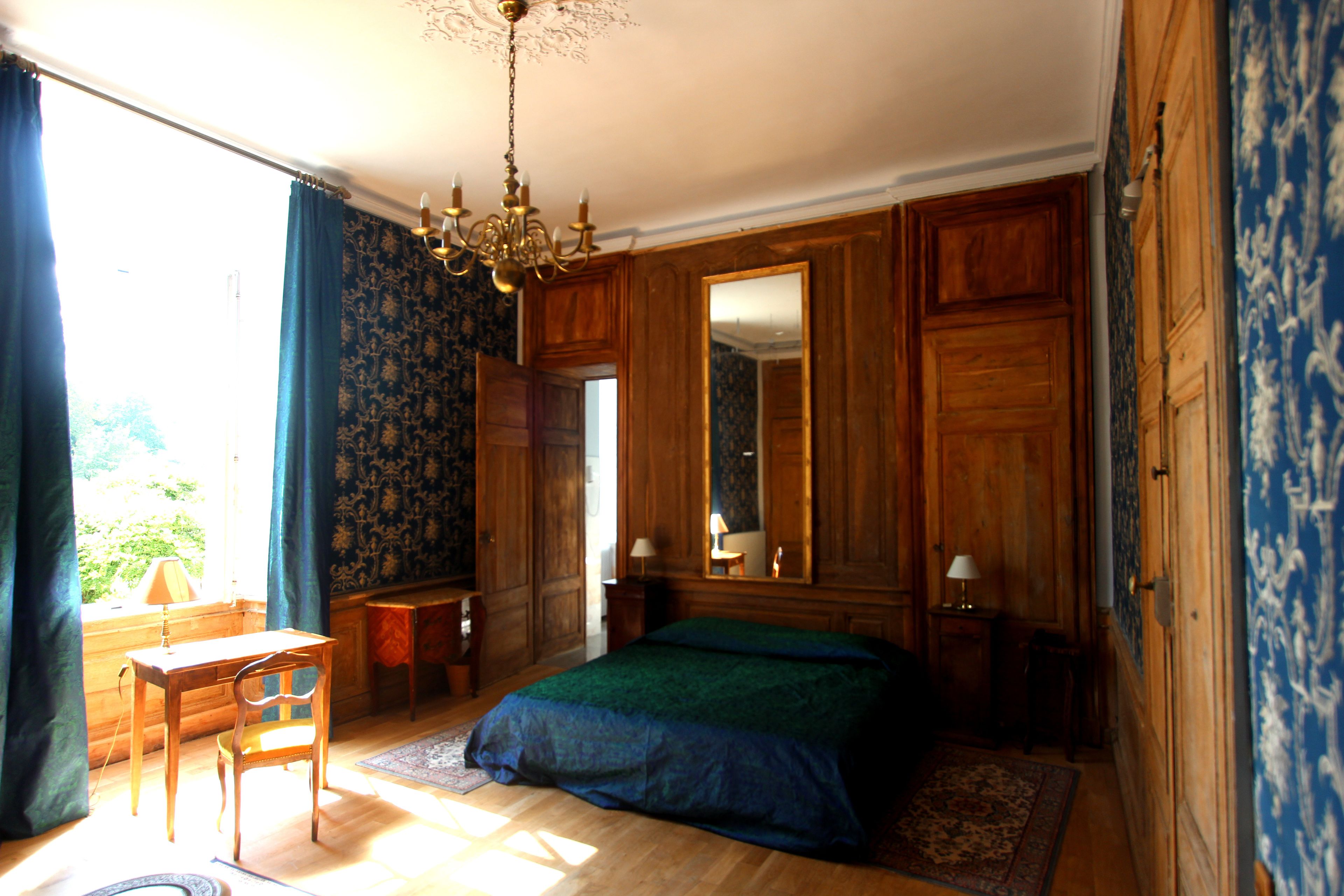 Un dormitorio con un armario de madera, una cama con colcha verde, un escritorio con silla y una lámpara de araña.