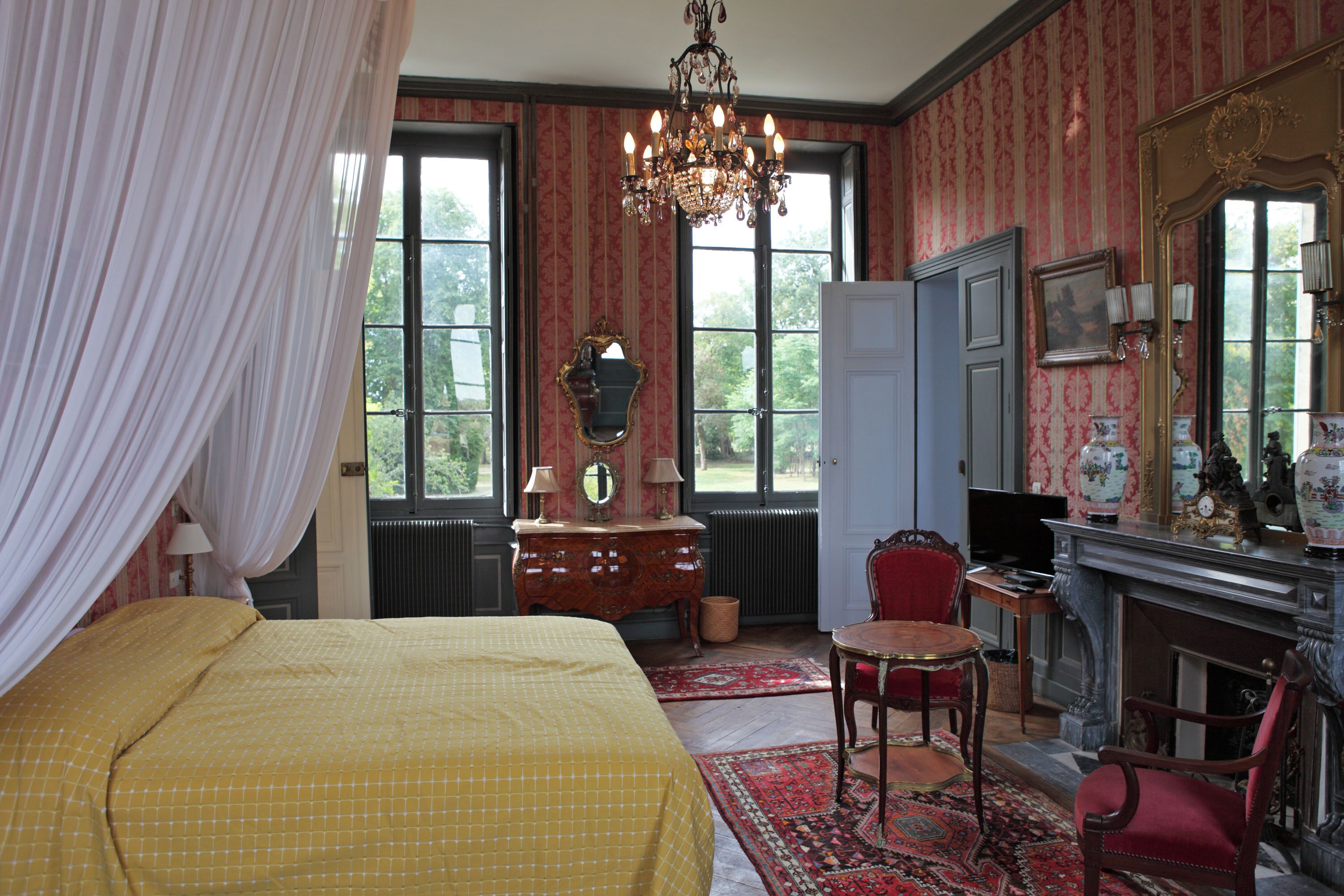 Una habitación con una cama, un sillón rojo, un aparador de madera, un espejo, una araña de cristal y una chimenea.