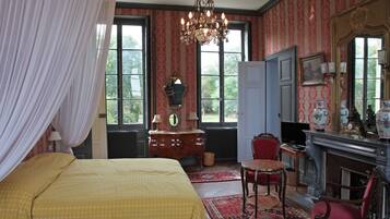 Una habitación con una cama, un sillón rojo, un aparador de madera, un espejo, una araña de cristal y una chimenea.